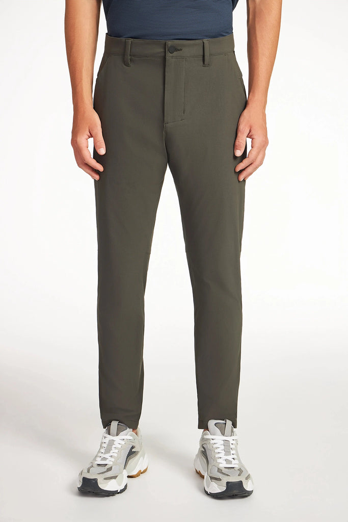 An individual stands against a white background wearing TECH APPAREL MENS DRI-FLEX PANTS in olive green, paired with a navy shirt and white-gray sneakers.