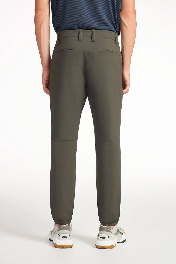 A person faces away from the camera wearing olive green TECH APPAREL MENS DRI-FLEX PANTS, a navy shirt, and light sneakers against a plain white background.