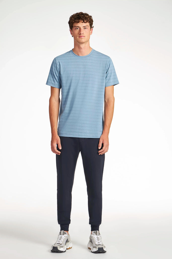 A young man stands facing forward against a plain light background, wearing a blue short-sleeve t-shirt, TECH APPAREL MENS AERO-KNIT JOGGER made from stretch fabric, and white sneakers.