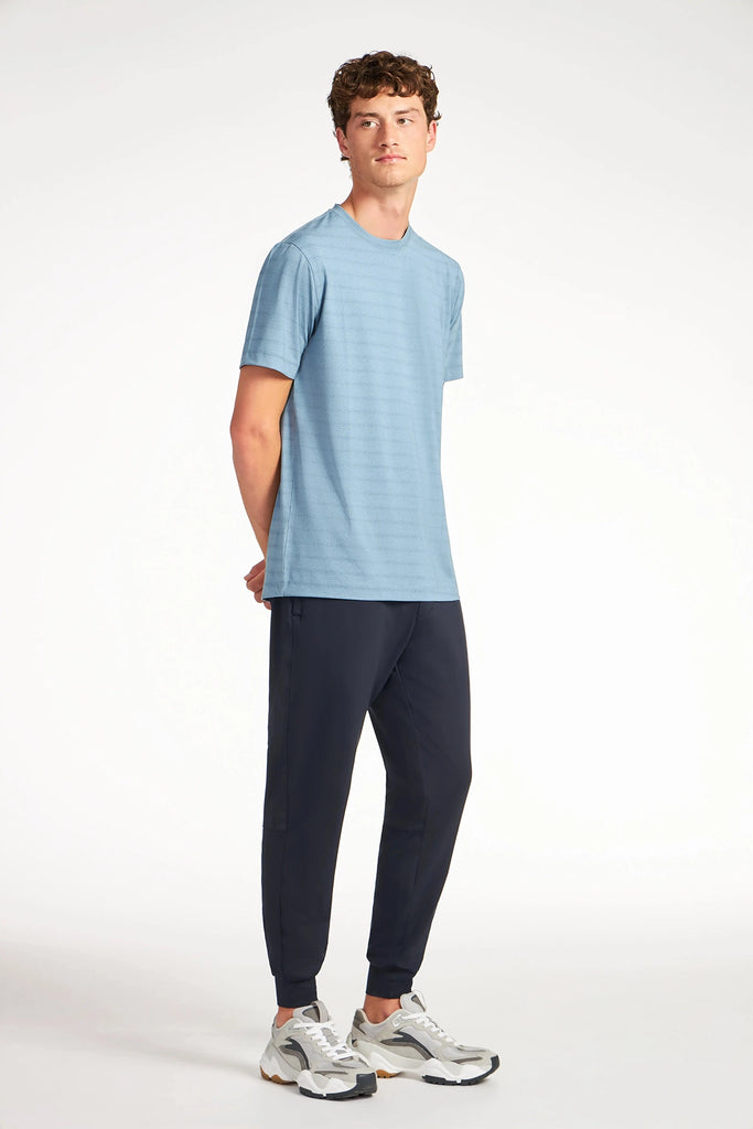 A young man stands against a white background, wearing a light blue short-sleeve t-shirt, white sneakers, and TECH APPAREL MENS AERO-KNIT JOGGER made from stretch fabric. He looks slightly to the side with his hands behind his back.