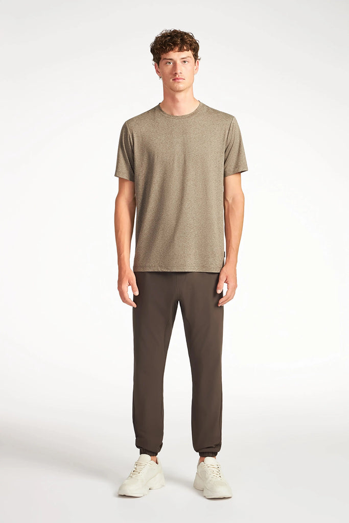 A young man faces forward, wearing a brownish-grey short-sleeve t-shirt, TECH APPAREL MENS AERO-KNIT PANTS made from stretch fabric, and white sneakers against a plain white background.
