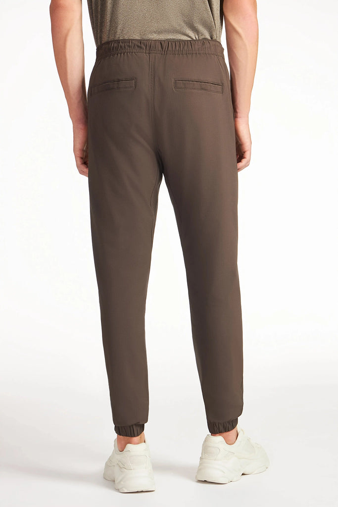 A man, seen from the back, wears brown TECH APPAREL MENS AERO-KNIT PANTS with white sneakers. The pants have stretch fabric, elastic waistband and cuffs, and two welt pockets. The plain background is light-colored.