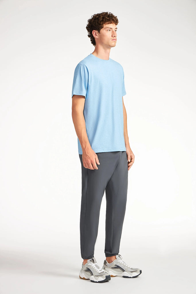 A young man with curly hair stands slightly turned, wearing a light blue t-shirt and TECH APPAREL MENS AERO-KNIT PANTS, featuring wrinkle-resistant, stretch fabric, plus gray sneakers, set against a plain white background.