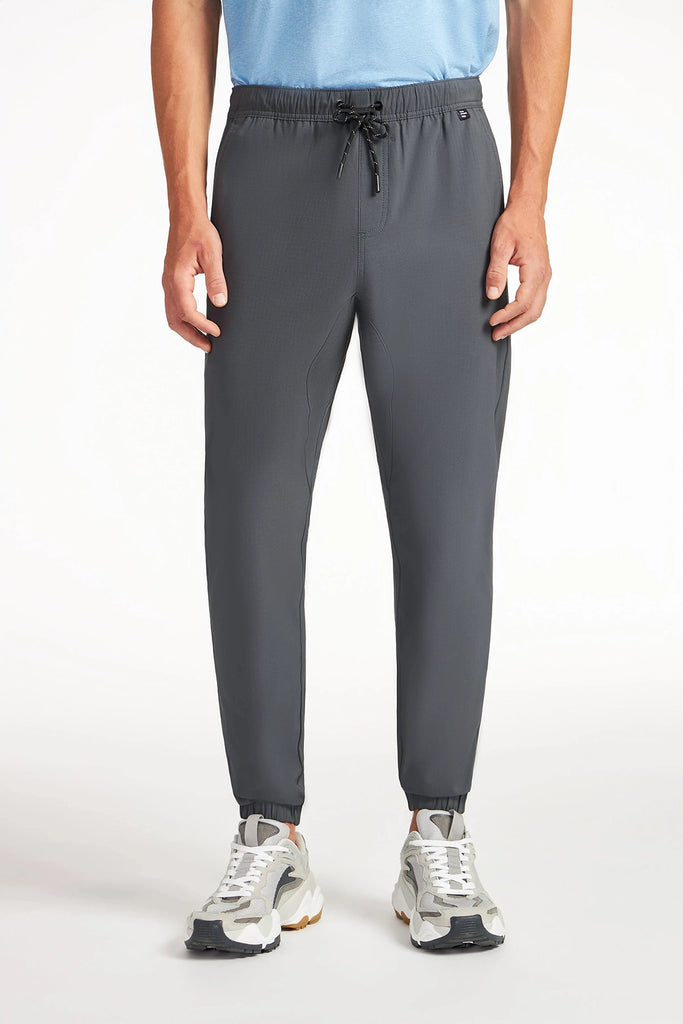 A person stands facing forward in TECH APPAREL MENS AERO-KNIT PANTS—gray, wrinkle-resistant stretch joggers with an elastic waistband and black drawstring—paired with a light blue shirt and white sneakers against a plain white background.