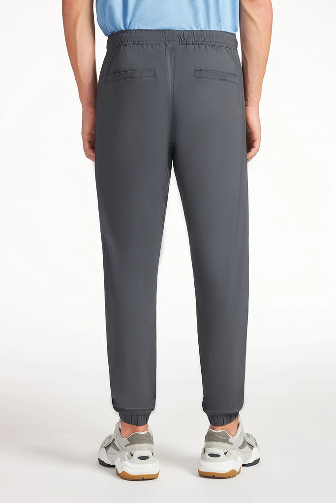 A man is seen from the back wearing TECH APPAREL MENS AERO-KNIT PANTS in dark gray, featuring stretch fabric, an elastic waistband, wrinkle-resistant cuffs, and two back welt pockets, paired with light-colored sneakers against a plain white background.