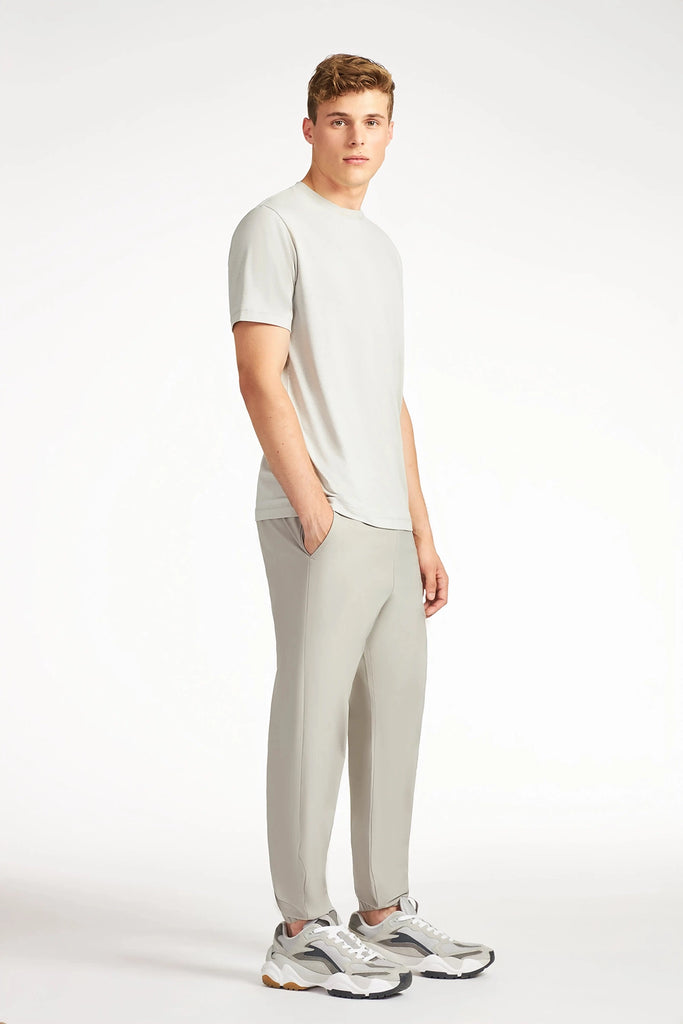 A young man with short hair stands against a white background wearing TECH APPAREL MENS AERO-KNIT PANTS, a matching tee, and chunky sneakers. The pants are stretchable and wrinkle-resistant. He gazes to the side with a neutral expression.