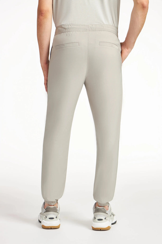 A person, seen from behind with hands on hips, wears light gray TECH APPAREL MENS AERO-KNIT PANTS—wrinkle-resistant joggers featuring elastic cuffs and two back pockets—paired with white sneakers.