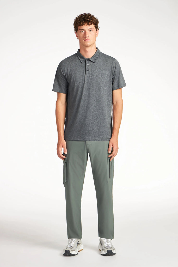 A young man stands facing forward against a white background, wearing TECH APPAREL’s MENS THERMAL FLEX UTILITY PANT in sage green, a charcoal gray polo shirt, and white sneakers.