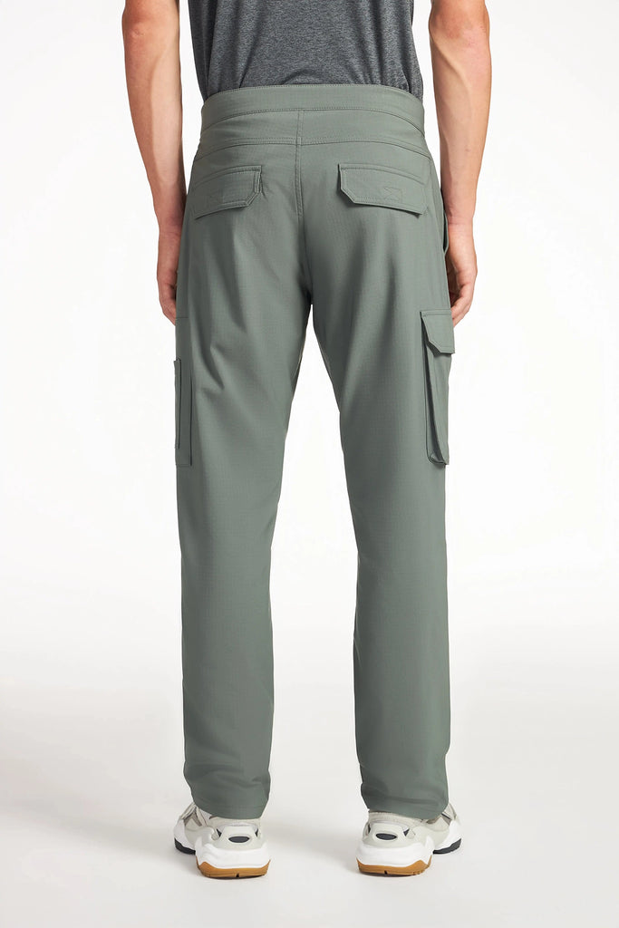 A person wearing TECH APPAREL MENS THERMAL FLEX UTILITY PANT in sage green, paired with a dark gray shirt and white sneakers, stands facing away from the camera against a plain white background.