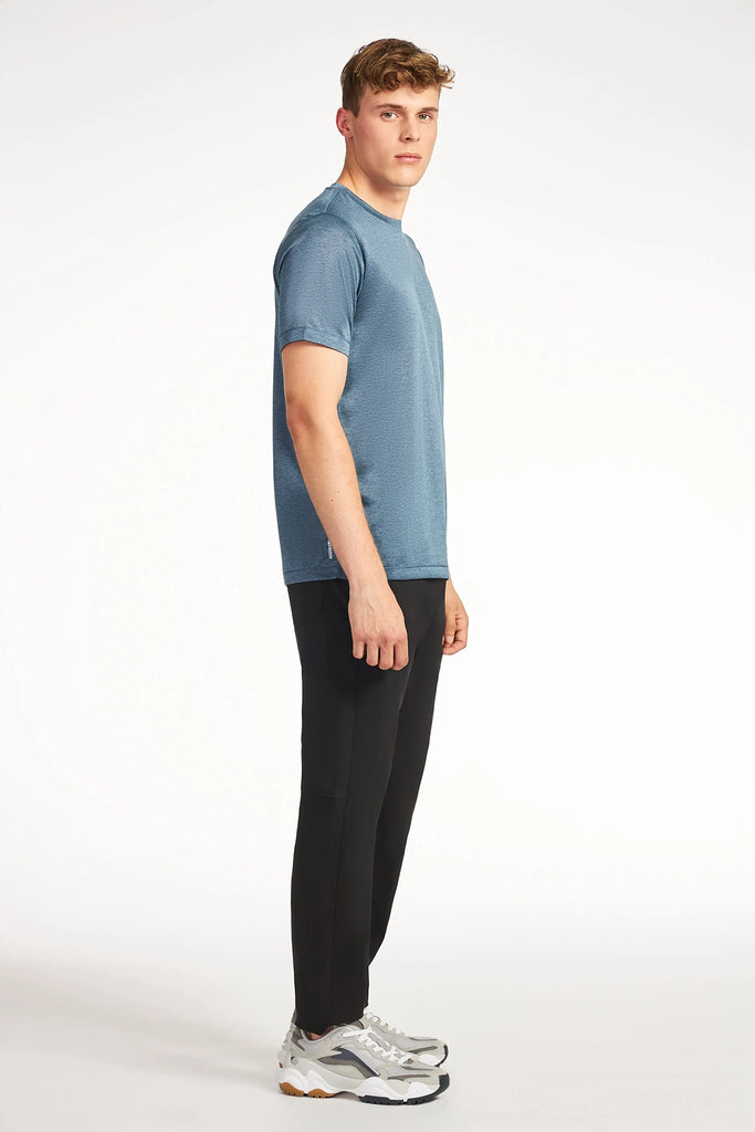 A young man stands sideways, looking at the camera, wearing a blue short-sleeve shirt, TECH APPAREL MENS COZY FLEEZE PANT, and white sneakers against a plain white background.