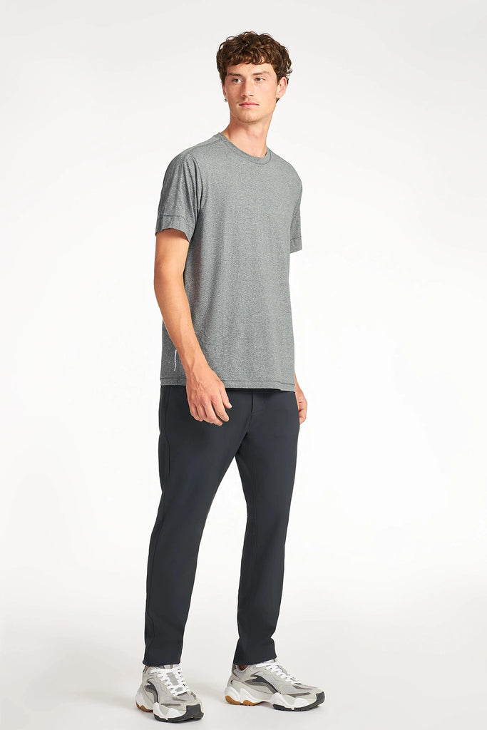 A young man stands against a plain background in TECH APPARELs MENS COZY FLEEZE PANT (navy jogger style), a gray t-shirt, and white athletic sneakers, looking slightly left with a neutral expression.