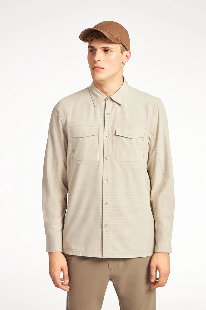A young man stands facing forward against a white background, wearing TECH APPARELs MENS FOUR WAY STRETCH SHIRT in light beige with breast pockets, along with brown pants and a brown cap.