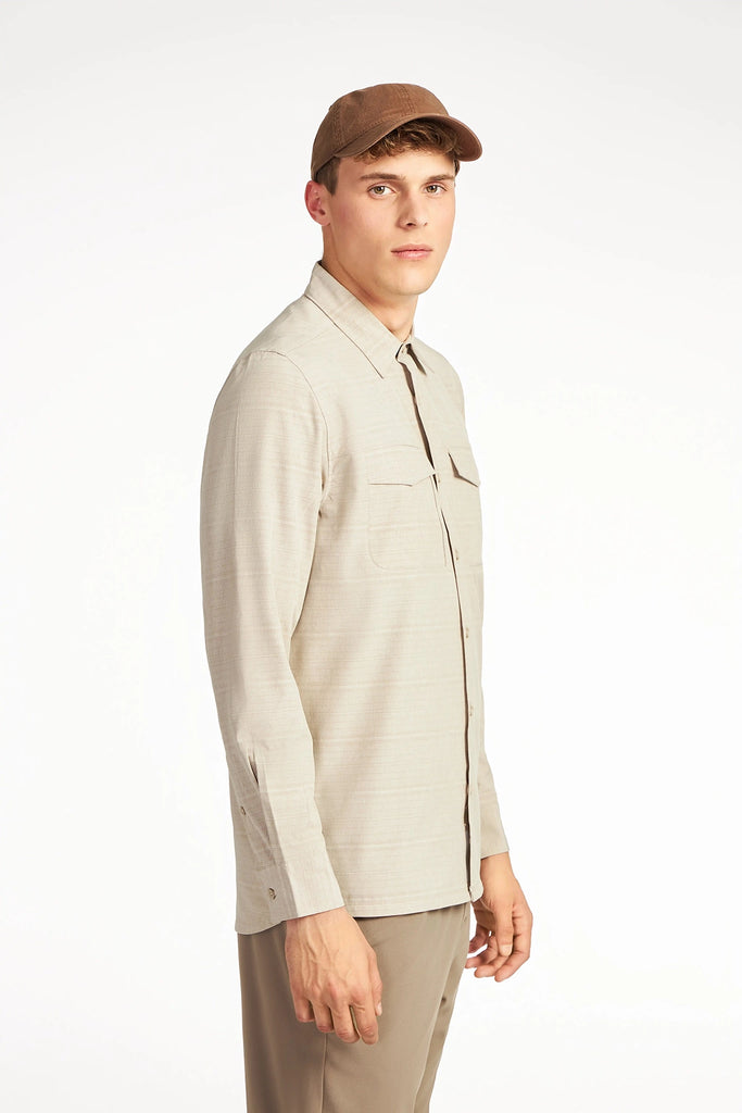 A young man in a TECH APPAREL MENS FOUR WAY STRETCH SHIRT and tan pants, with a brown cap, stands against a white background, looking at the camera with a neutral expression.