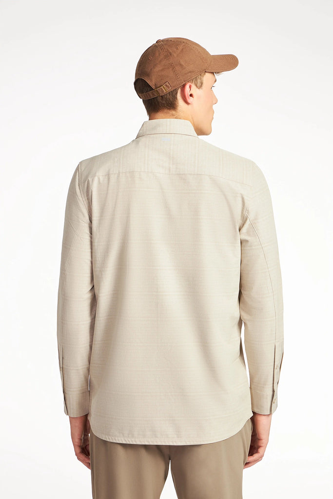 A man stands with his back to the camera against a white background, wearing TECH APPAREL’s MENS FOUR WAY STRETCH SHIRT in beige, paired with brown pants and a brown baseball cap.