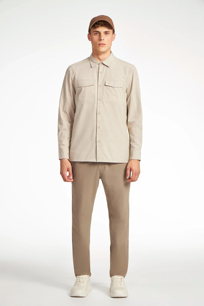 A man faces forward in a studio, wearing the TECH APPAREL MENS FOUR WAY STRETCH SHIRT in light beige with two chest pockets, matching beige pants, white sneakers, and a brown cap against a plain white background.