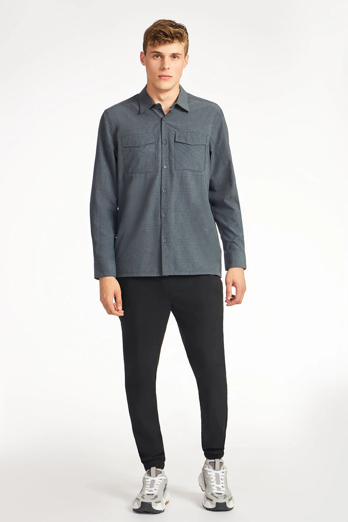 A young man faces forward, wearing TECH APPARELs MENS FOUR WAY STRETCH SHIRT in blue, paired with black pants and white-gray sneakers, all set against a plain white background.