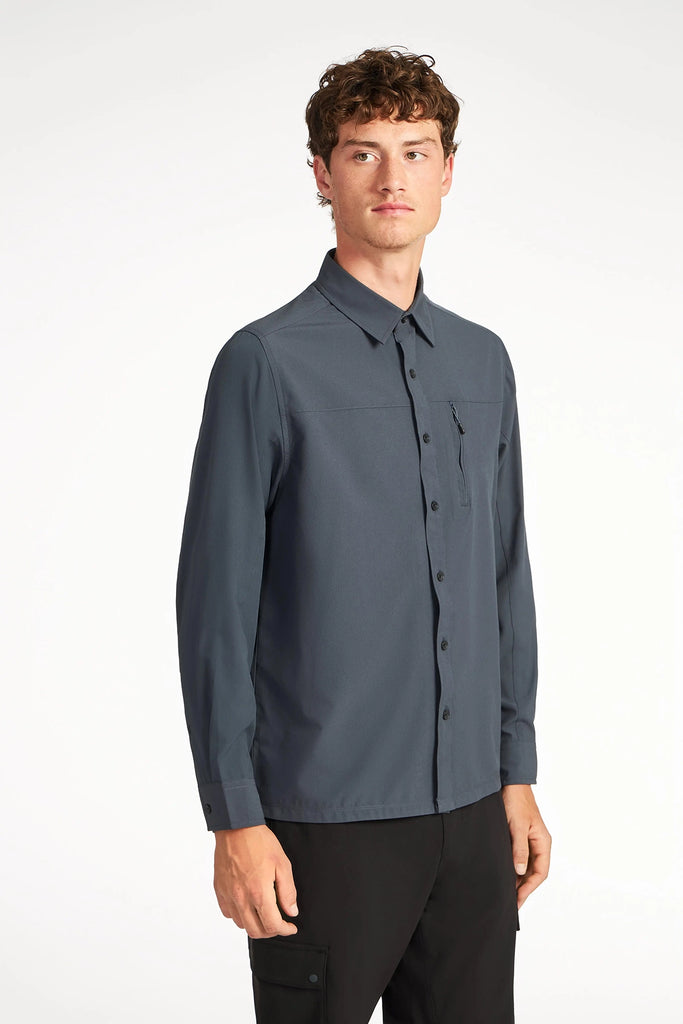 A young man with short curly hair models the TECH APPAREL MENS SOLID STRETCH RIBSTOP SHIRT in dark gray, featuring a chest pocket and breathable, comfortable fabric, standing against a plain white background.