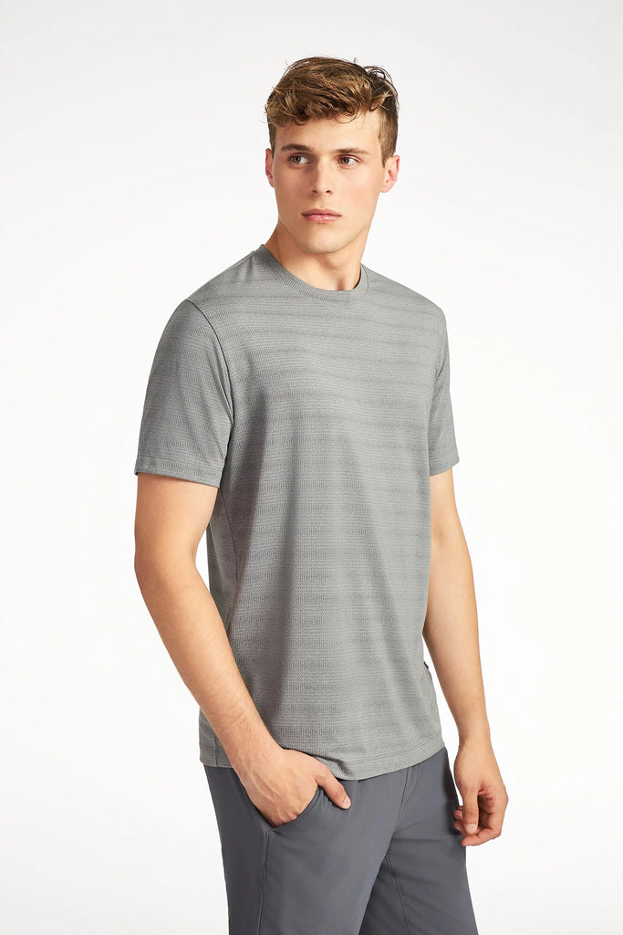 A young man with short, light brown hair wears TECH APPARELs MENS MIX JACQUARD STRIPE T-SHIRT in light grey and dark grey pants, standing against a plain white background and looking slightly to his left.