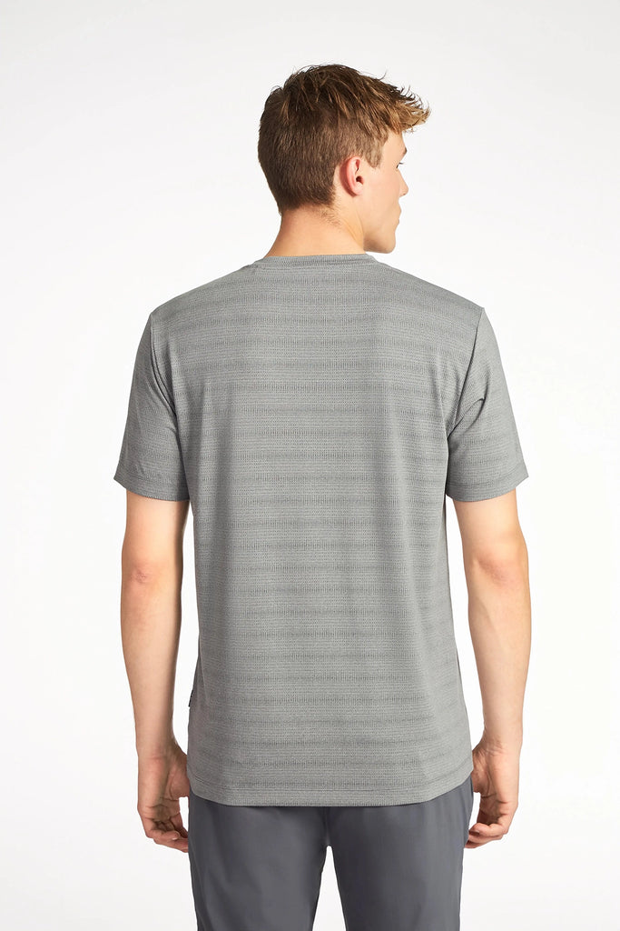 A man with short brown hair stands with his back to the camera, wearing the TECH APPAREL MENS MIX JACQUARD STRIPE T-SHIRT and dark gray pants—an essential modern wardrobe look—against a plain white background.