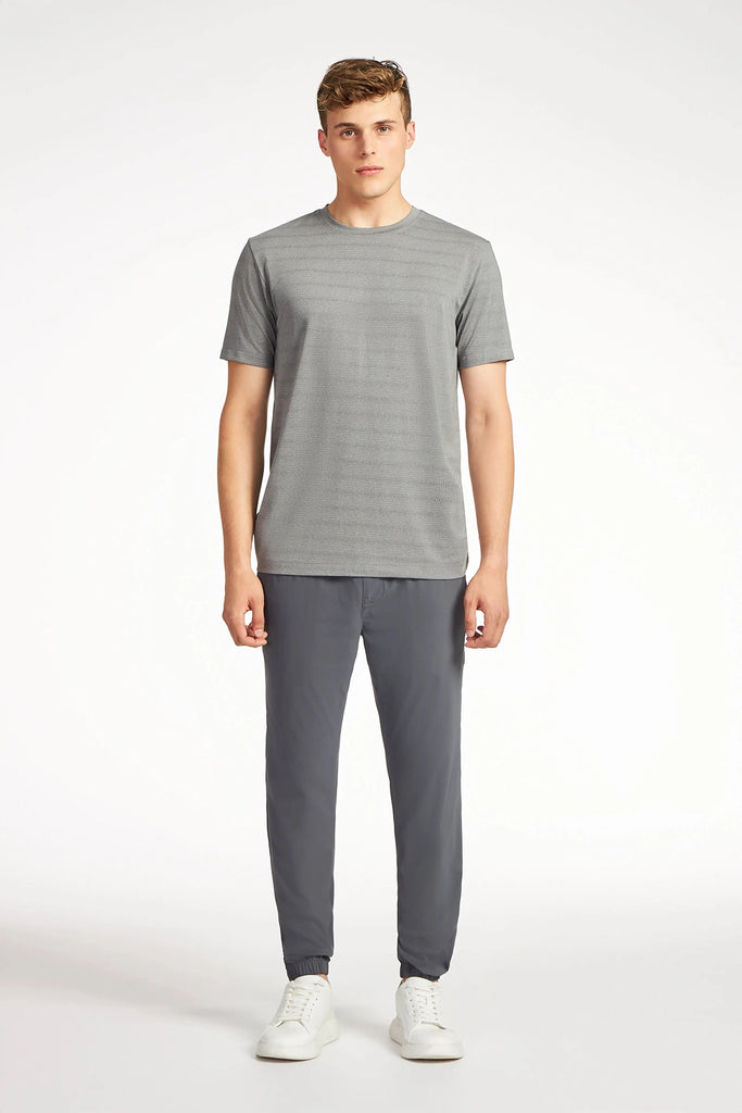 A young man stands facing forward in TECH APPAREL’s MENS MIX JACQUARD STRIPE T-SHIRT, paired with dark gray joggers and white sneakers—timeless pieces for any men’s wardrobe—against a plain white background.
