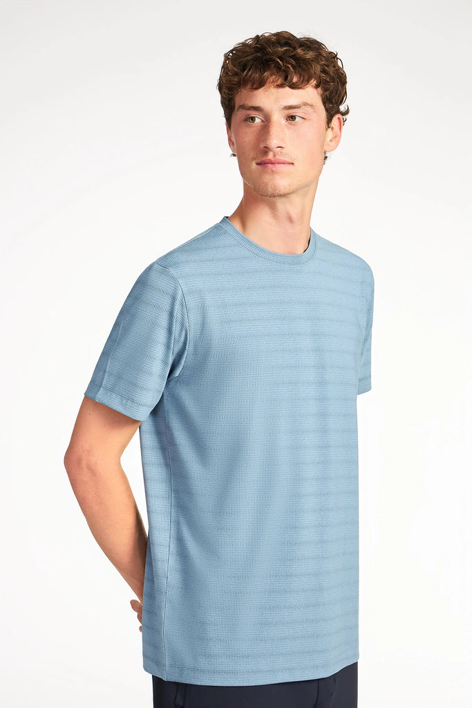 A young man with short curly hair wears the TECH APPAREL MENS MIX JACQUARD STRIPE T-SHIRT and dark pants, standing against a plain white background while looking slightly to the side.