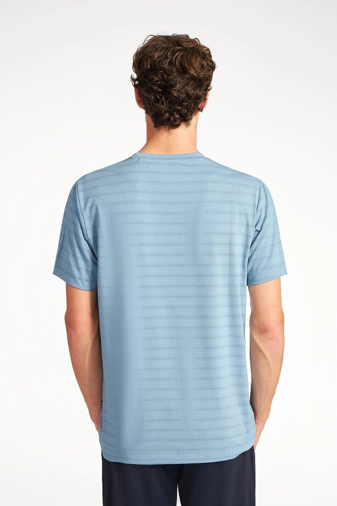 A person with short, curly brown hair is shown from the back, wearing a light blue TECH APPAREL MENS MIX JACQUARD STRIPE T-SHIRT and dark pants against a plain white background.