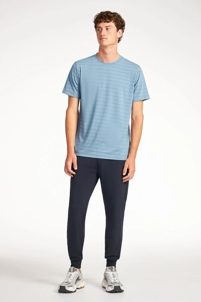 A young man stands against a white background, wearing the TECH APPAREL MENS MIX JACQUARD STRIPE T-SHIRT in light blue with dark jogger pants and white sneakers. He looks slightly to the side with a neutral expression.
