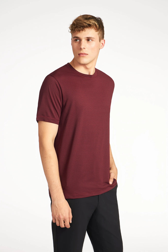 A young man with short, light brown hair wears a TECH APPAREL MENS VARIEGATED STRIPE CREWNECK T-SHIRT and black pants, standing against a white background and looking to his left with one hand in his pocket.