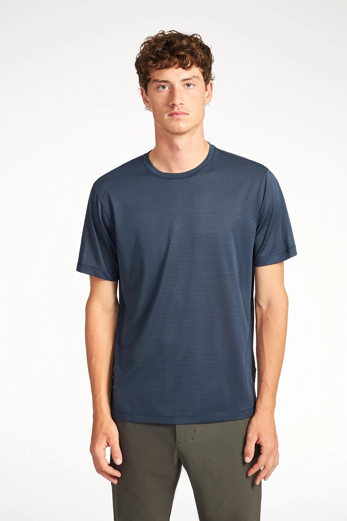 A young man with short curly brown hair wears the TECH APPAREL MENS VARIEGATED STRIPE CREWNECK T-SHIRT and olive green pants, standing against a white background and looking straight at the camera.