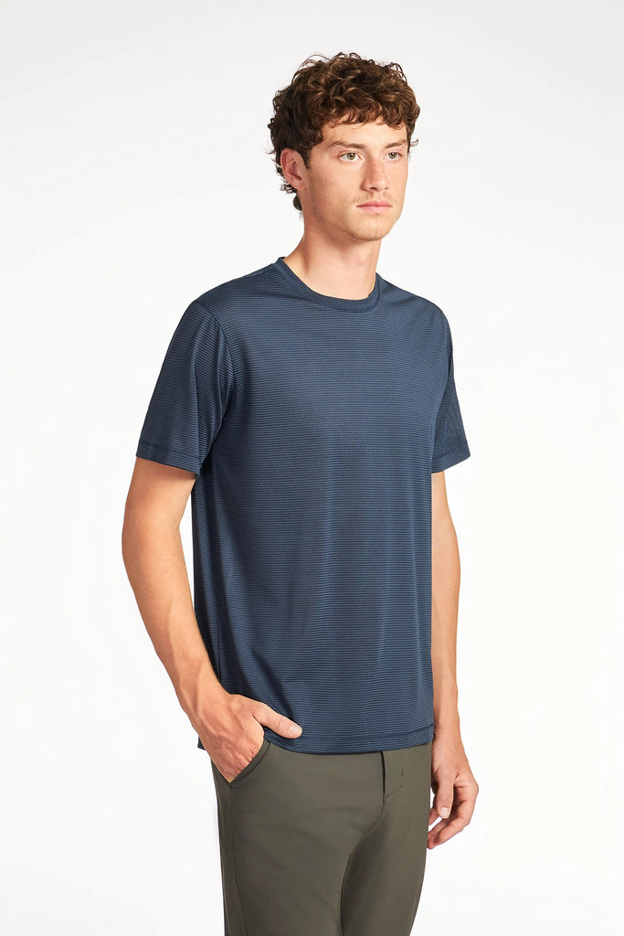 A young man with short, curly brown hair models the TECH APPAREL MENS VARIEGATED STRIPE CREWNECK T-SHIRT and olive green pants, posing against a plain white background with one hand in his pocket.