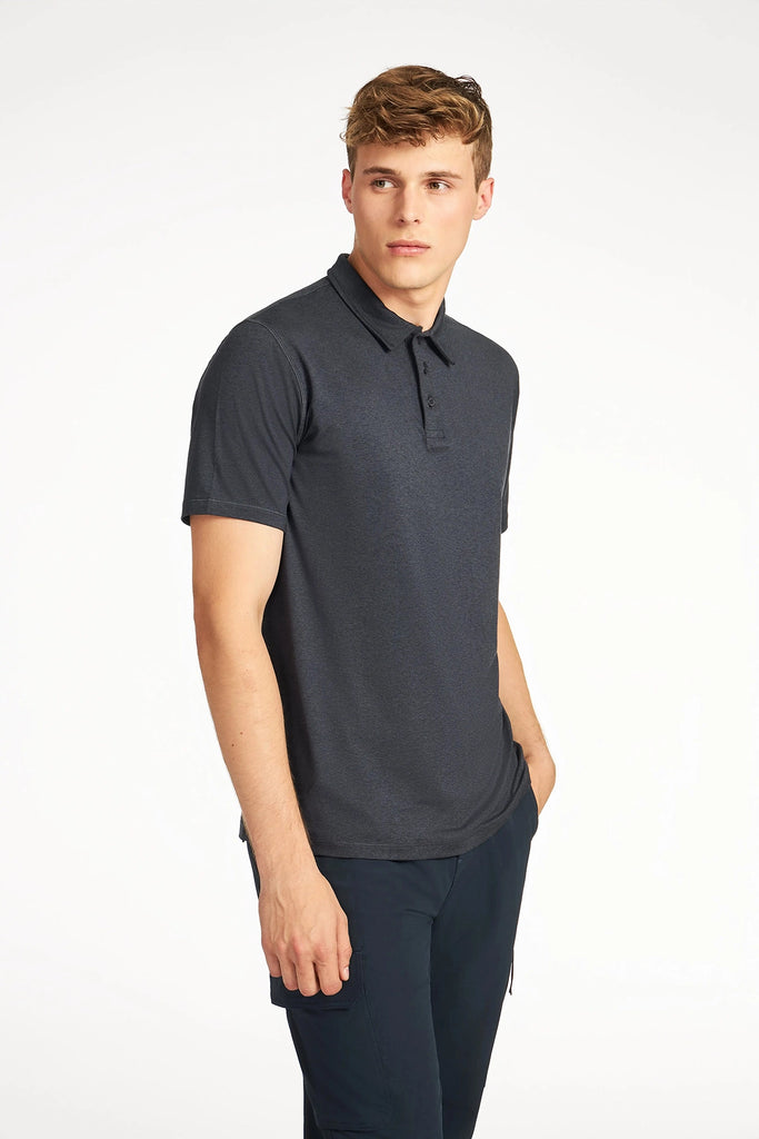 A young man with short blond hair wears the TECH APPAREL MENS BRUSHED SPACE DYED POLO and dark pants, standing with one hand in his pocket and looking slightly to the side against a plain white background.