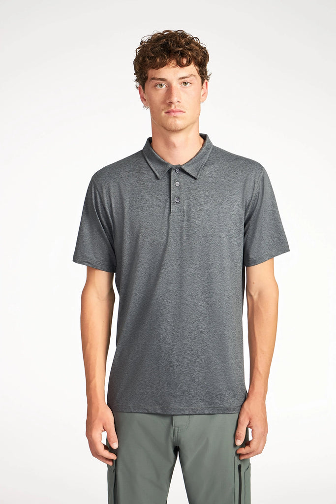 A young man with short curly hair models the TECH APPAREL MENS BRUSHED SPACE DYED POLO in dark gray and gray pants, standing against a white backdrop. The polos wrinkle-resistant fabric ensures a sharp look all day.