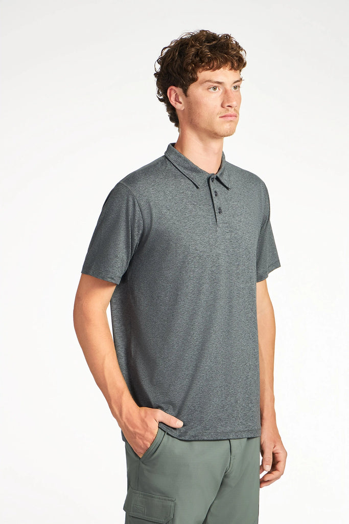 A young man with curly brown hair wears the TECH APPAREL MENS BRUSHED SPACE DYED POLO and green cargo pants, standing against a plain white background with one hand in his pocket, looking slightly to the side.