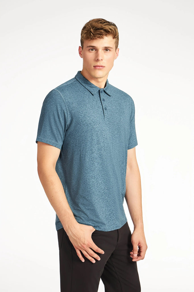 A young man with short, light brown hair wears the TECH APPAREL MENS BRUSHED SPACE DYED POLO and black pants, standing against a plain white background and looking directly at the camera.