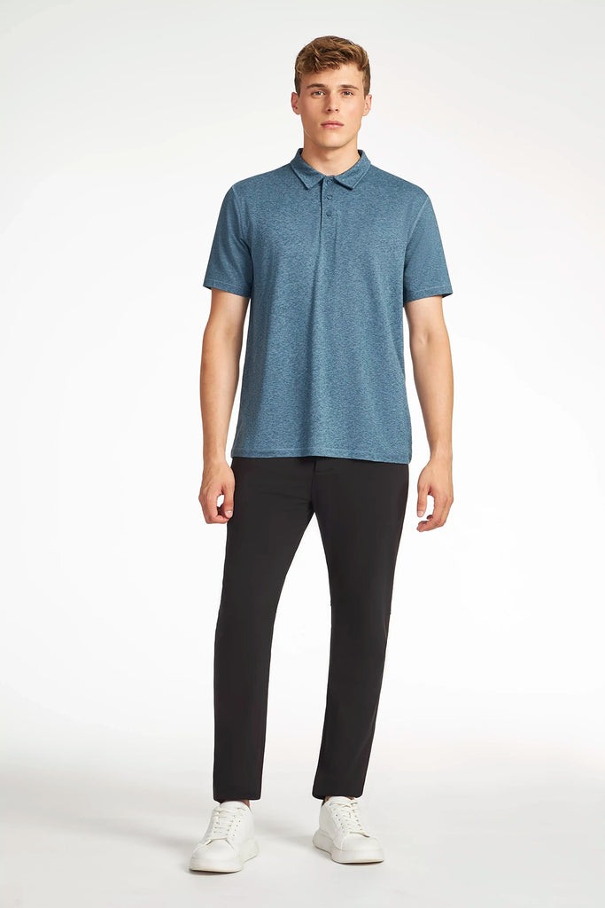 A young man faces forward in a TECH APPAREL MENS BRUSHED SPACE DYED POLO (blue), black pants, and white sneakers—an ideal look set against a plain white background.