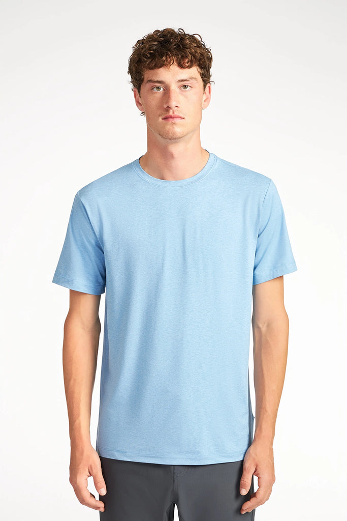 A young man with short curly hair wears the TECH APPAREL MENS BRUSHED SPACE DYED T-SHIRT in light blue and dark pants, standing facing forward against a plain light background with a neutral expression.