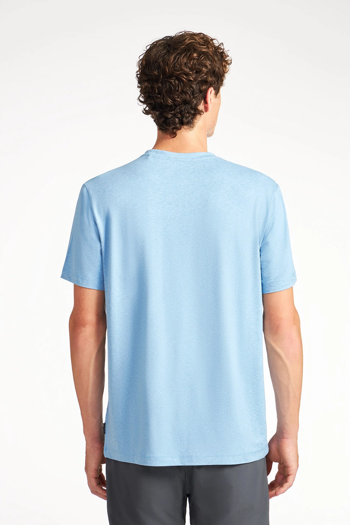 A person with short curly brown hair is seen from behind against a plain white background, wearing a light blue TECH APPAREL MENS BRUSHED SPACE DYED T-SHIRT and dark gray shorts.