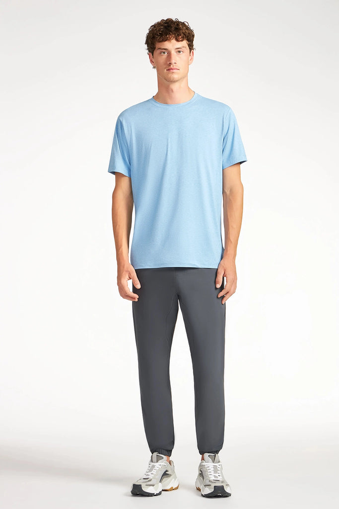 A person stands facing forward, wearing the TECH APPAREL MENS BRUSHED SPACE DYED T-SHIRT in light blue, dark gray jogger pants, and white sneakers against a plain white background.
