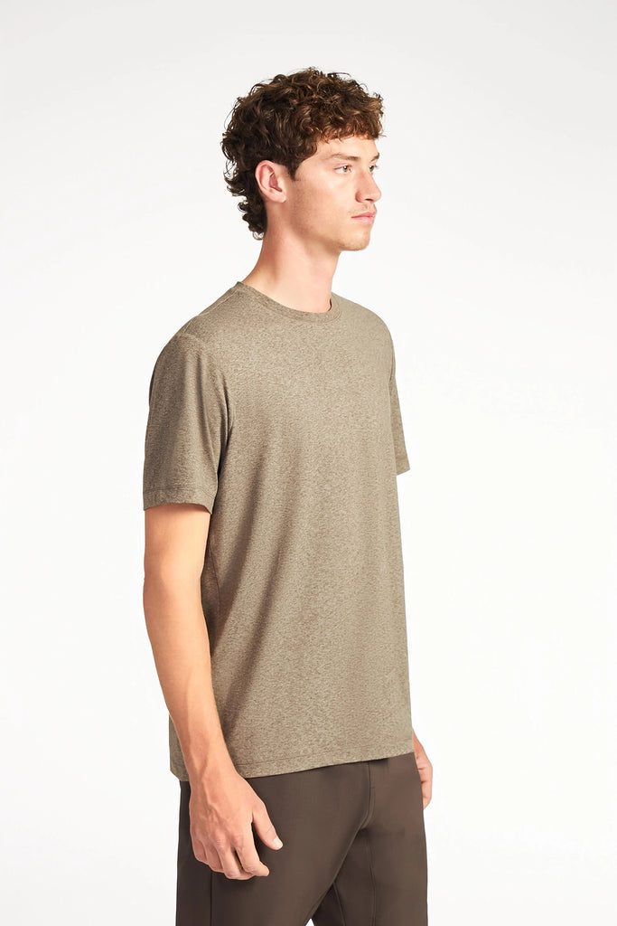 A young man with curly brown hair stands sideways against a plain white background, wearing TECH APPARELs wrinkle-resistant MENS BRUSHED SPACE DYED T-SHIRT in light brown and dark brown pants.