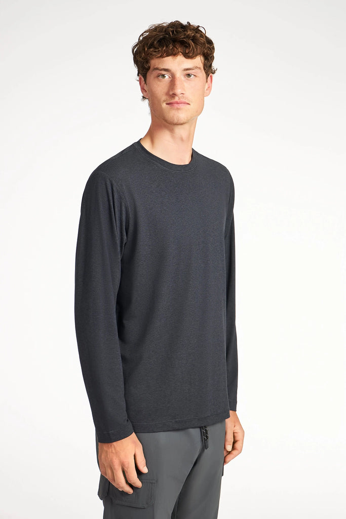 A young man with short curly hair models the TECH APPAREL MENS BRUSHED SPACE DYE LONG SLEEVE TOP and gray pants, standing slightly to the side against a plain white background.