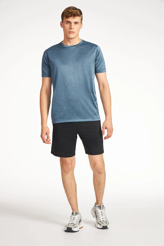 A young man stands against a white background wearing the TECH APPAREL MENS MARLED PIQUEE T-SHIRT in blue, paired with black shorts and gray sneakers. He gazes forward with a neutral expression, arms relaxed at his sides.