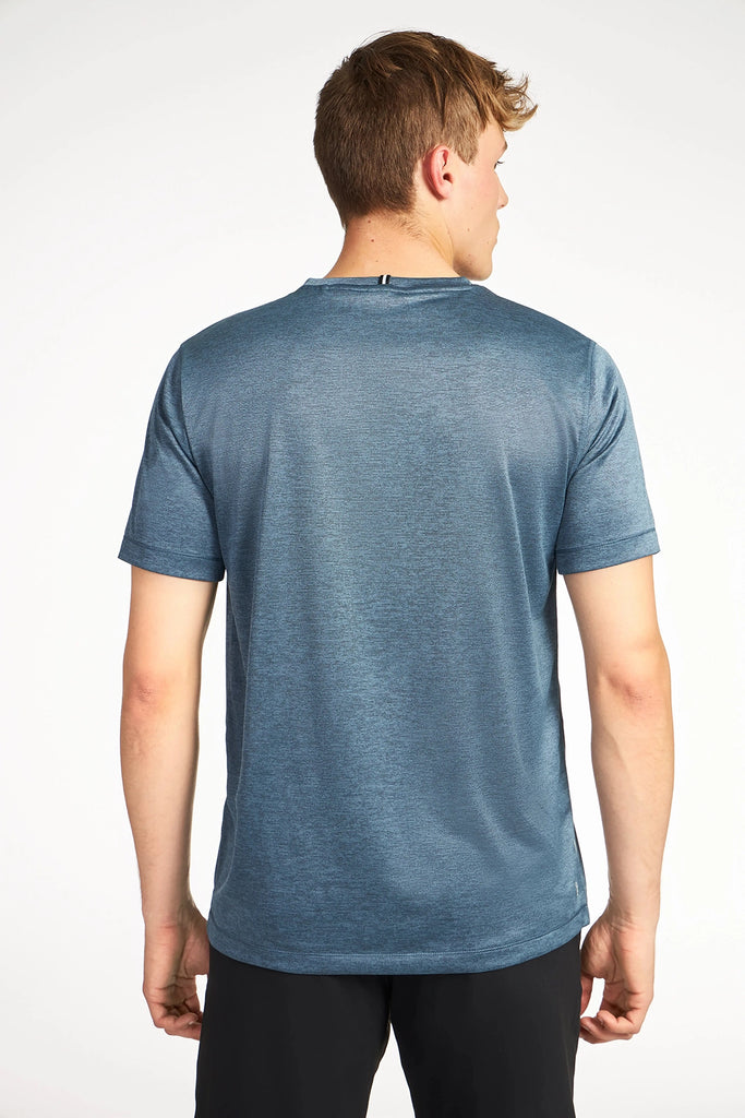 A young man with short light brown hair is seen from the back, wearing TECH APPARELs MENS MARLED PIQUEE T-SHIRT and black shorts against a plain white background.