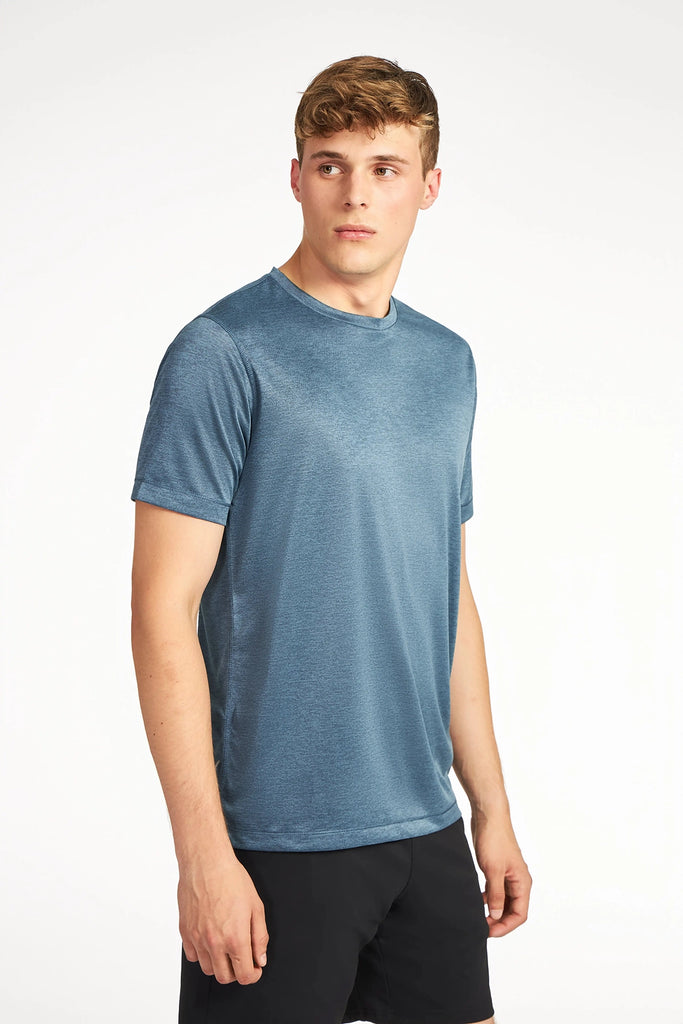 A young man with short light brown hair wears a blue TECH APPAREL MENS MARLED PIQUEE T-SHIRT made of stretch fabric and black shorts, standing against a plain white background and looking slightly to his left.