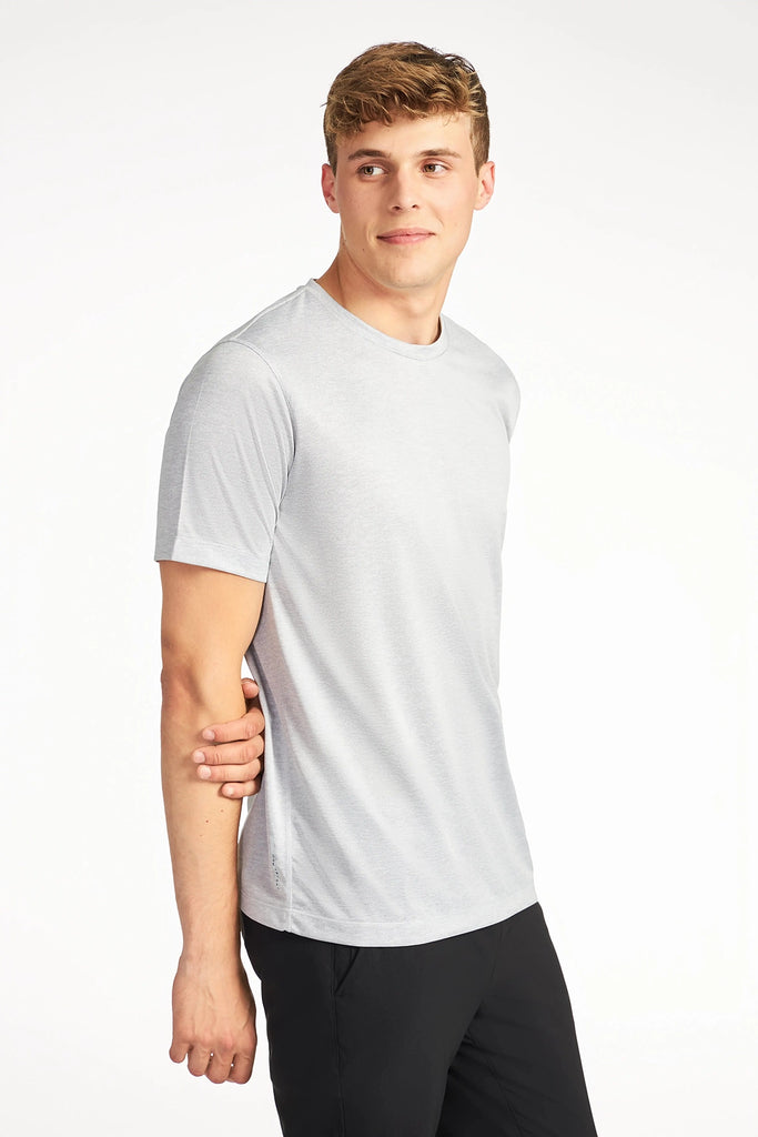 A young man wears the TECH APPAREL MENS MARLED PIQUEE T-SHIRT in light gray and black pants, standing slightly sideways with a hand resting on his arm. The background is plain white.