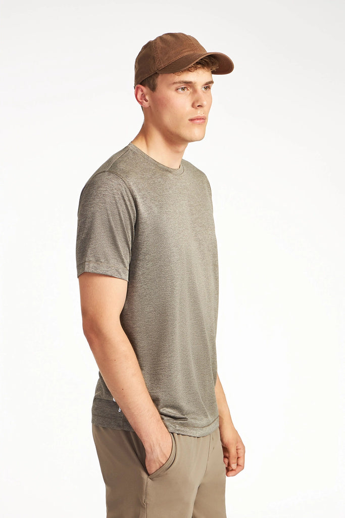A young man in a TECH APPAREL MENS MARLED PIQUEE T-SHIRT, brown cap, and beige pants stands with hands in pockets, gazing to the side against a plain white background.