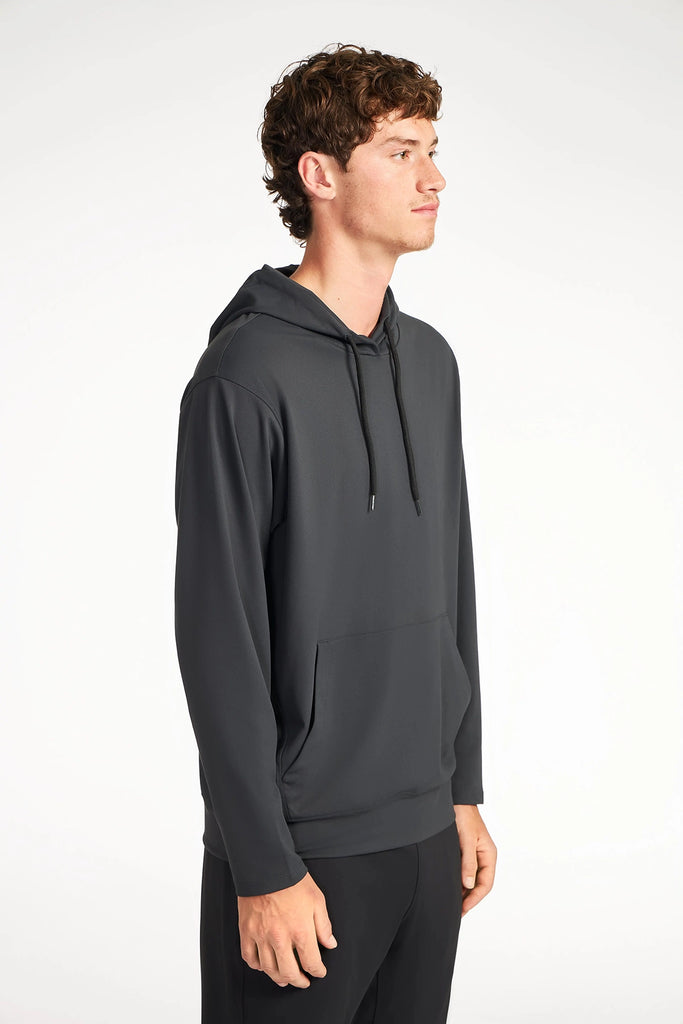 A young man with short curly brown hair stands in profile against a white background, wearing the TECH APPAREL MENS TECH:SOFT HOODIE in dark gray, featuring active fabric, a front pocket, and drawstrings.