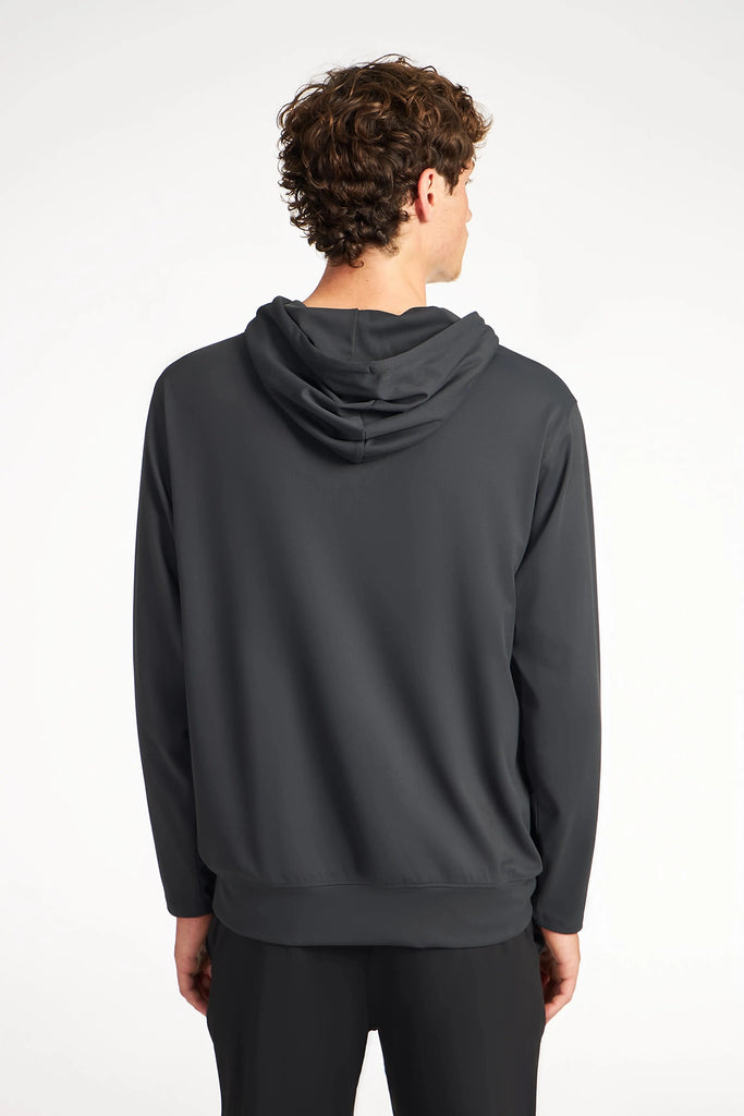 A person with curly hair faces away from the camera, wearing a TECH APPAREL MENS TECH:SOFT HOODIE in dark gray and black pants against a plain light background.