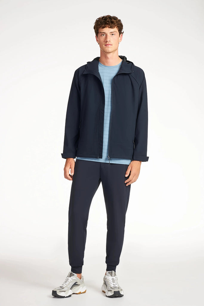 A young man faces forward in a TECH APPAREL MENS DRI-FLEX JACKET, light blue tee, navy joggers, and white sneakers. This lightweight jacket combines comfort and style against a plain white background.