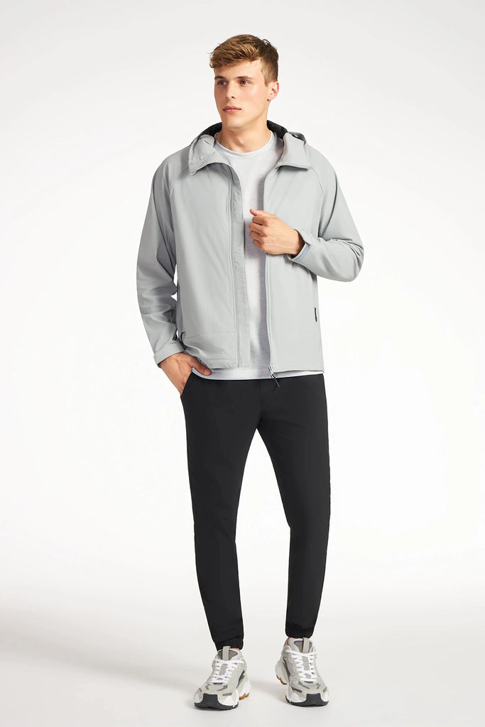 A man stands against a white background wearing TECH APPAREL’s MENS DRI-FLEX JACKET, a white t-shirt, black pants, and gray and white sneakers. This lightweight jacket offers breathable comfort; he poses with one hand in his pocket.
