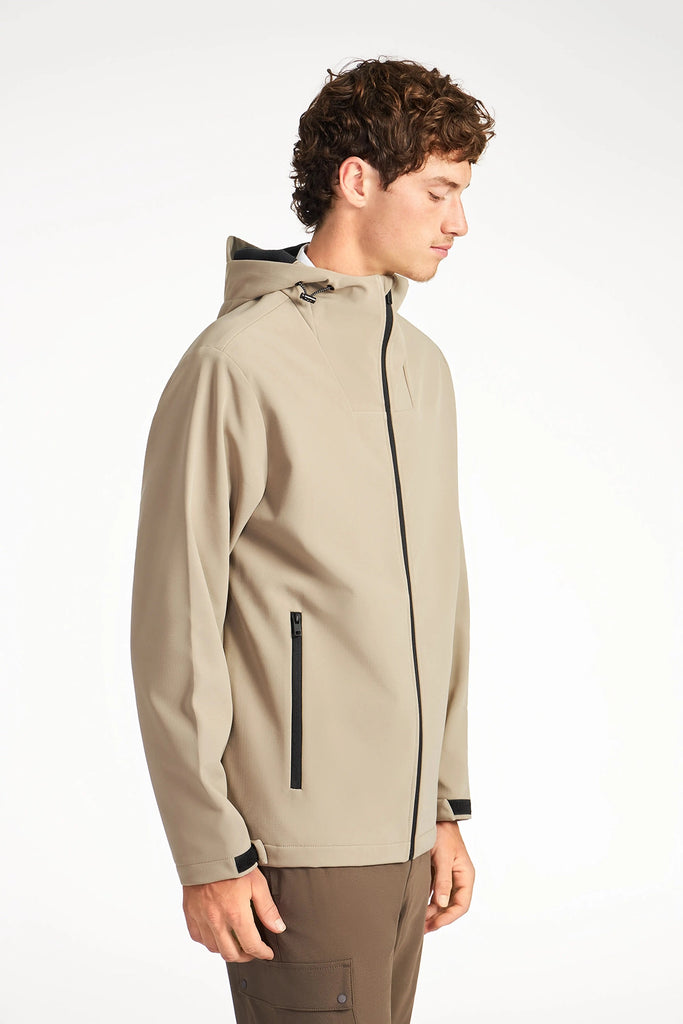 A young man with curly hair is wearing a light beige TECH APPAREL MENS BONDED FLEECE JACKET with black zippers and dark pants, standing sideways against a plain white background.